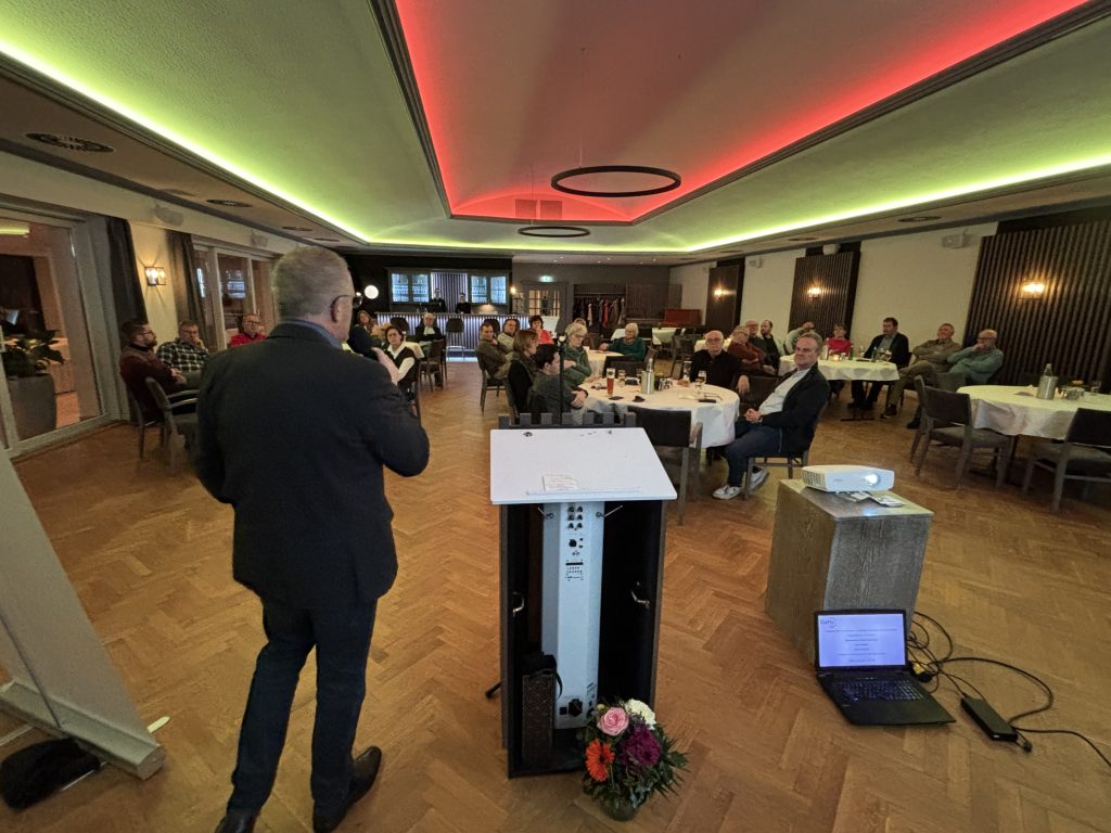 Ein Mann steht in einem Saal, in dem etwa 40 Personen an Tischen sitzen. Er spricht in ein Mikrofon.