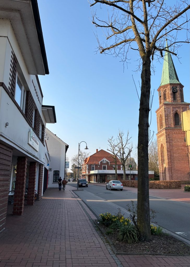 Straße entlang einer Kirche auf der rechten Seite. Links von der Straße eine Parkbucht vor einer Häuserzeile.