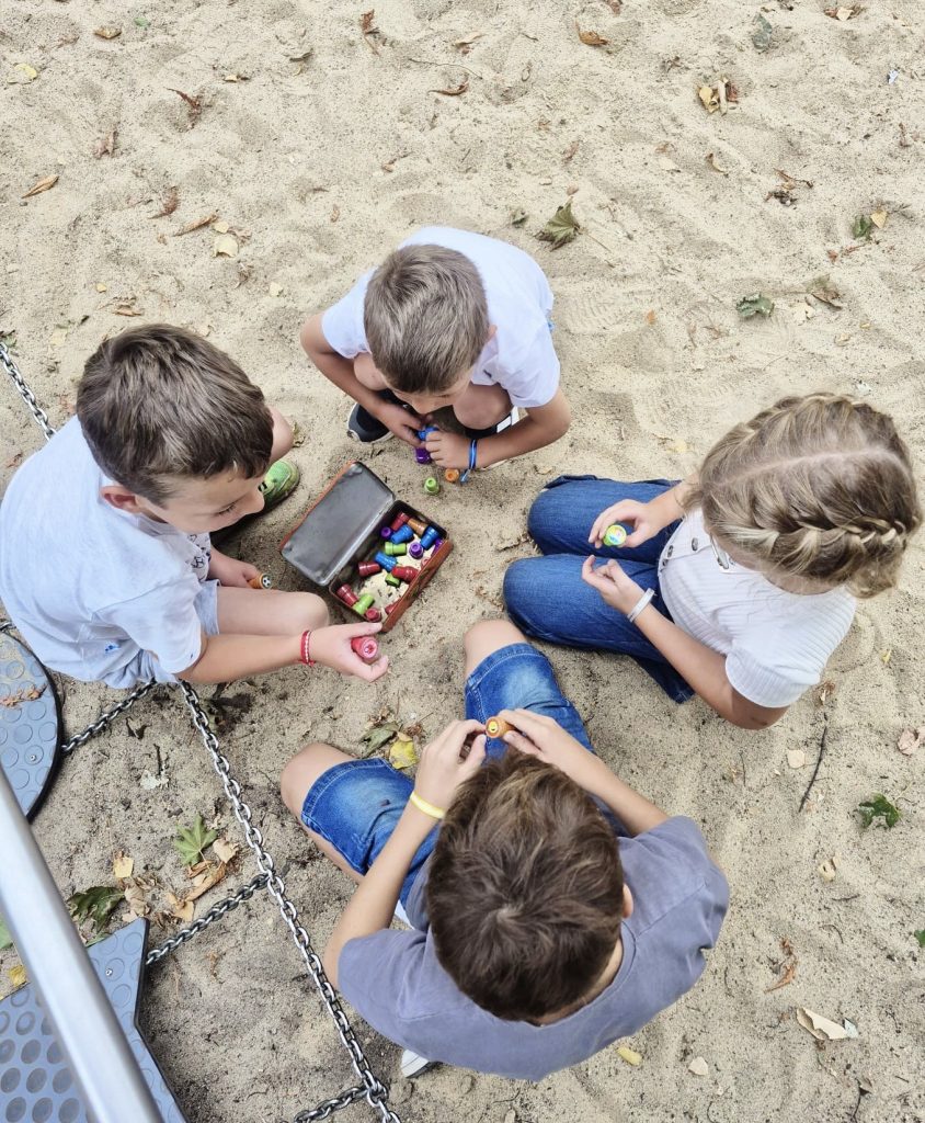 Blick von oben auf vier Kinder, die auf einer Sandspielfläche um eine kleine, bunt gefüllte Schatzkiste herum knien.