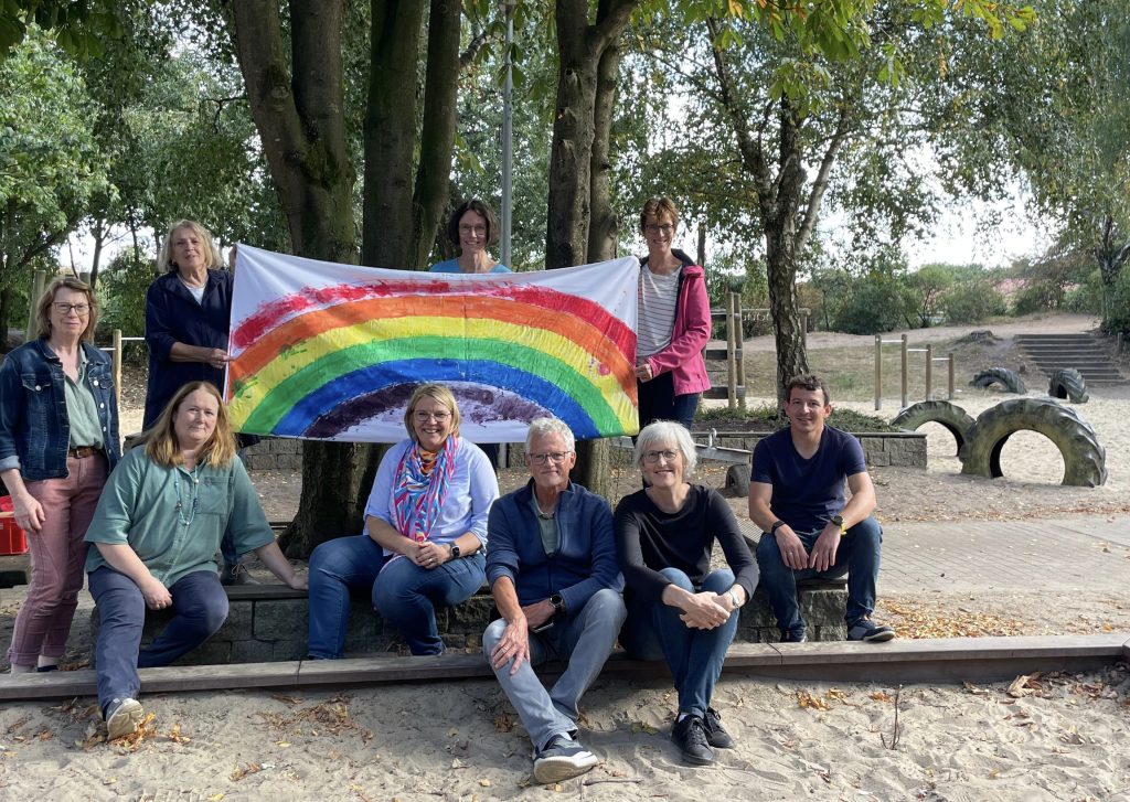 Neun Erwachsene stehen und sitzen lächelnd um ein Regenbogentransparent.