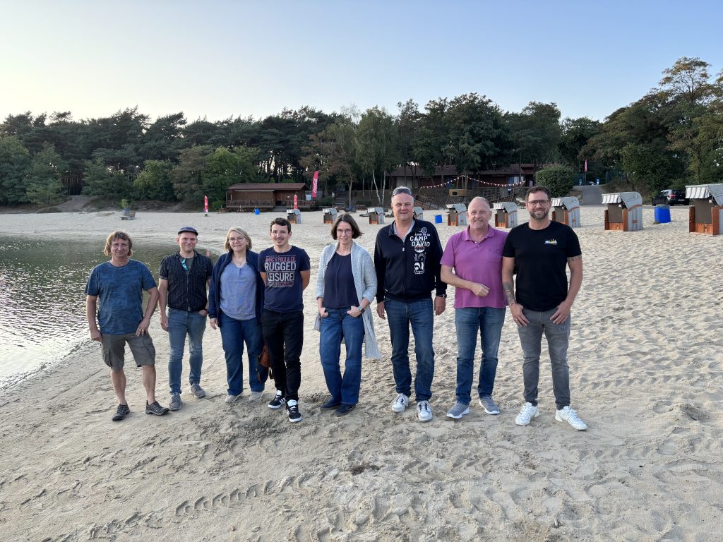 Acht Personen stehen nebeneinander am Sandstrand eines Badesees. Im Hintergrund Bäume und ein Spielplatz.