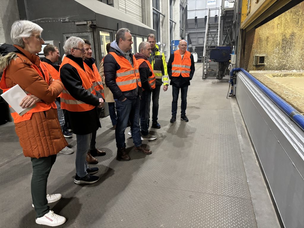 Eine Grupe von etwa zehn Personen in orangen Warnwesten steht in einer Industriehalle links vom Rand einer großen Wanne. 